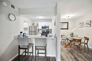 A modern kitchen with white cabinets, a stainless steel refrigerator, and a microwave. There is a dining area with a table and chairs. - I041 – New 2BD Apt | Pool | DFW South 183/161 (Irving)
