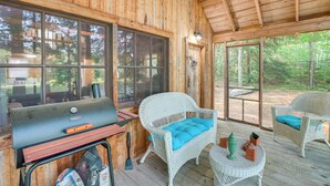 Eine Holzhütte mit großem Fenster, Grill, Korbmöbeln und Blick auf den Wald.