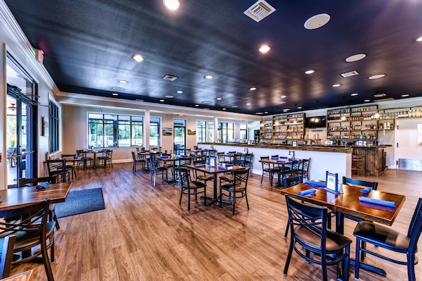 A spacious restaurant with wooden tables and chairs, a bar area, and large windows allowing natural light.