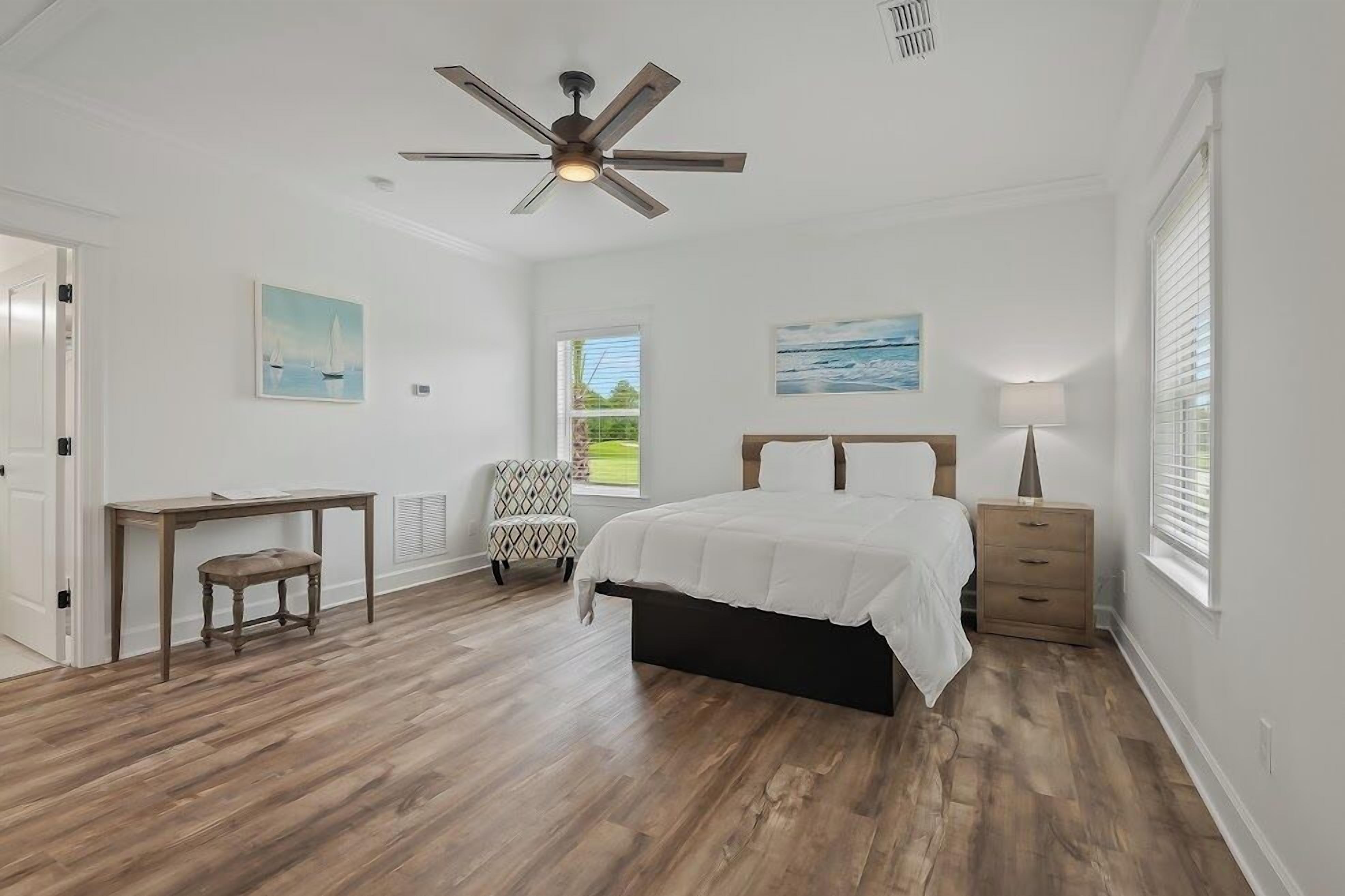 A bedroom with a bed, a desk, a chair, a ceiling fan, and a window.