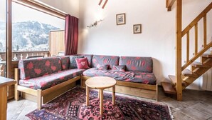 Un salon chaleureux avec un canapé en bois, une petite table ronde et une vue sur les montagnes par une grande fenêtre.