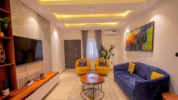 A modern living room with a flat-screen TV, a blue sofa, two yellow armchairs, a coffee table, and a large abstract painting. - BnJ Benin City Oasis - Spacious 3BR Apartment (Benin City)