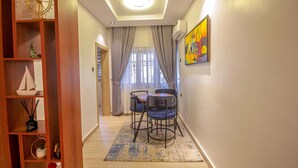 A modern living room with a wooden bookshelf, a dining area with a table and chairs, a large window with curtains, and a colorful abstract painting on the wall. - BnJ Benin City Oasis - Spacious 3BR Apartment (Benin City)