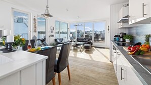 A modern kitchen with white cabinetry, a dining area with black chairs, and a large window offering a city view. - Sea Breeze: Meerblick, Sauna, Dachterrasse, Bootsanleger (Olpenitz)