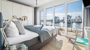 A modern bedroom with a large bed, a desk, and a view of the city and water. - Sea Breeze: Meerblick, Sauna, Dachterrasse, Bootsanleger (Olpenitz)