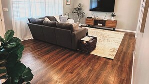 A modern living room with a grey sofa, wooden flooring, a flat-screen TV, and a large plant. - Gorgeous Florida Pool and Spa Home (Jacksonville)