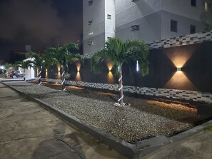 A nighttime view of a building with a tiled facade, a gravel pathway, and palm trees.