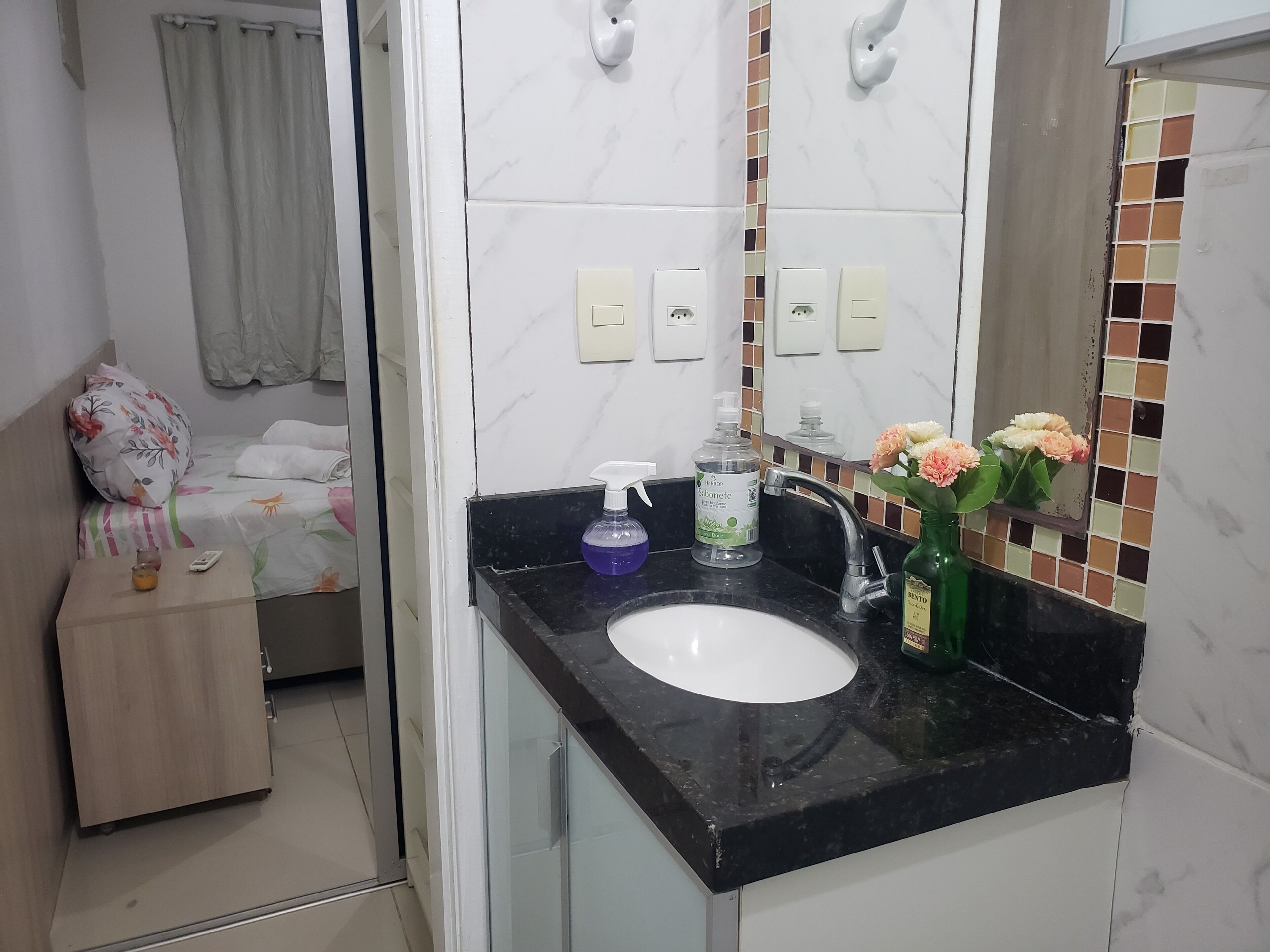 Une salle de bain avec un lavabo blanc, un plan de toilette noir et un miroir avec un pare-éclaboussures carrelé. On trouve des articles de toilette sur le plan de toilette, notamment un distributeur de savon et un flacon de gel hydroalcoolique.