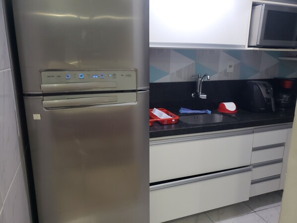 A stainless steel refrigerator with a control panel, a microwave above it, a sink with a faucet, and a red and white kettle on the countertop.