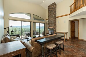 A spacious living area with a large stone fireplace, a dining table with bar stools, and a view of the mountains through large windows.
