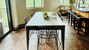 A spacious dining area with a long table, chairs, and a chandelier. Large windows offer views of the outdoors.