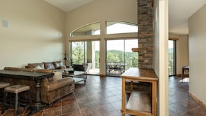 A spacious living room with a high ceiling, a stone fireplace, a dining area with a table and chairs, and large windows offering a view of the outdoors.