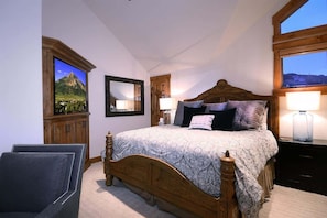 Um quarto com cama de madeira, duas mesas de cabeceira com abajures, uma cadeira, um espelho e uma janela com vista.