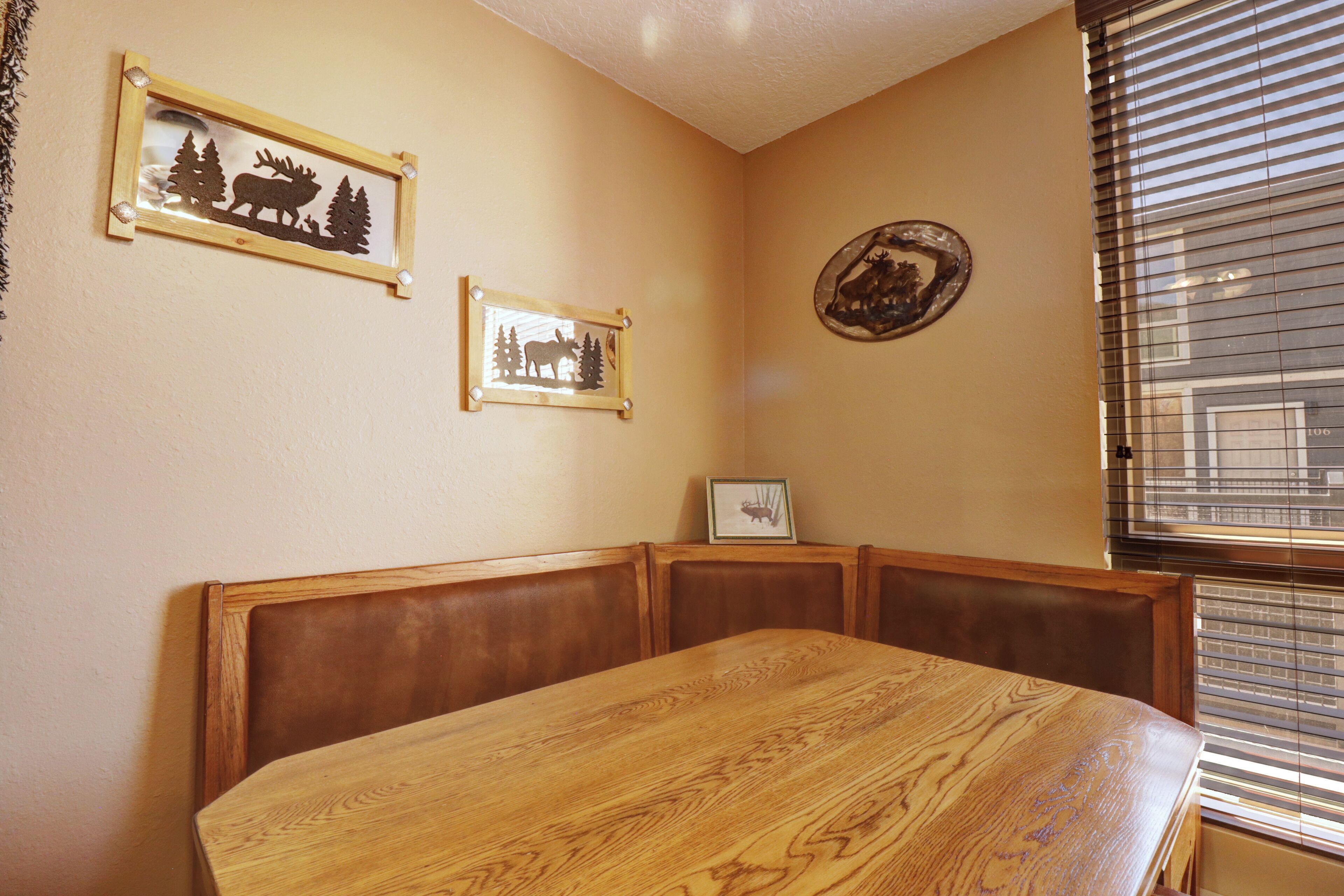 A dining area with a wooden table and booth seating, framed pictures on the wall, and a window with blinds.