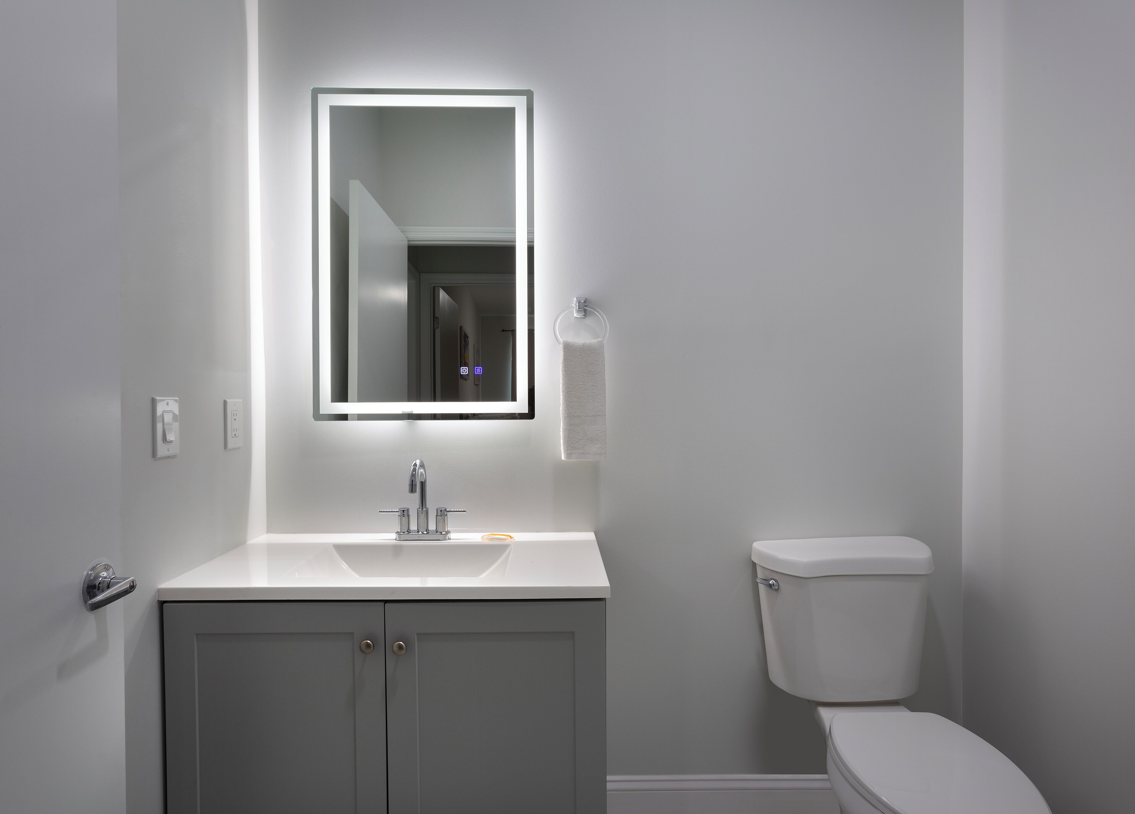 Un baño moderno con tocador blanco, espejo con iluminación integrada, inodoro y puerta con manija.