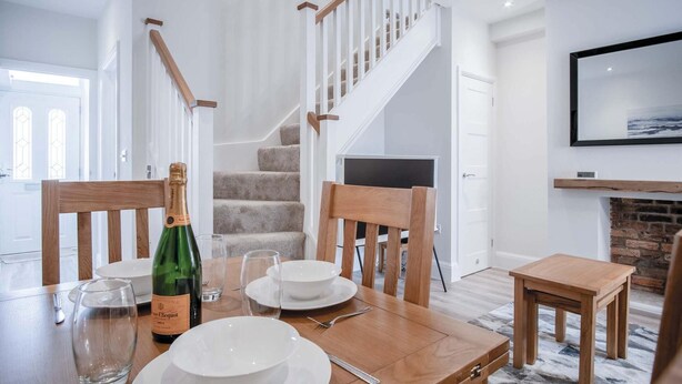 A dining area with a wooden table set for four, a bottle of champagne, and a television mounted on the wall. - Fleet Street - 2 Bedroom Holiday Home - Swansea (Swansea)