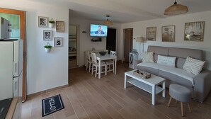 A modern living room with a sofa, coffee table, and TV. There is a dining area with chairs and a table. The room has a door, a wall-mounted TV, and a ceiling light.