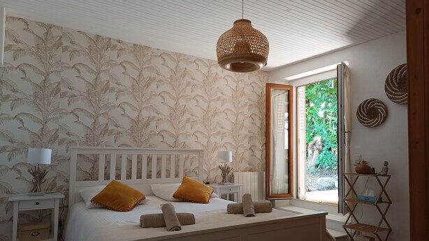 A bedroom with a bed, nightstands, a wooden headboard, a hanging light fixture, and a view of greenery outside.