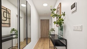 A hallway with a wooden floor, a black console table with a vase of green plants, and a large mirror on the left.