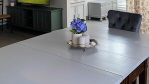 Uma área de refeições com uma mesa branca, uma cadeira estofada preta, um lustre, uma televisão sobre um móvel e vista para o exterior através de uma janela.