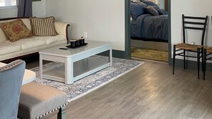Sala de estar com sofá, mesa de centro e cadeiras. Há um ventilador de teto, uma janela com cortinas e um quadro na parede.