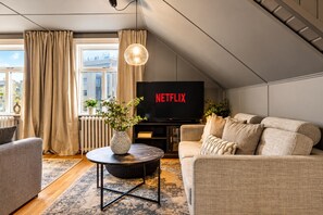 A living room with a sofa, a coffee table with a vase, a television displaying 'NETFLIX,' and a window with curtains.