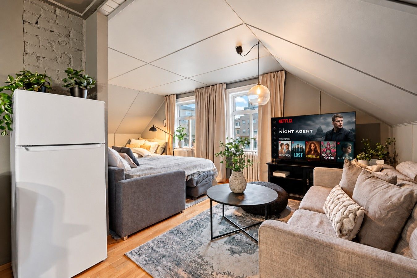 A modern loft bedroom with a bed, a sofa, a coffee table, and a TV displaying 'The Night Agent'.