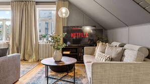 A cozy living room with a large flat-screen TV displaying 'NETFLIX', a comfortable sofa, a coffee table with a vase, and a view of the city through large windows.