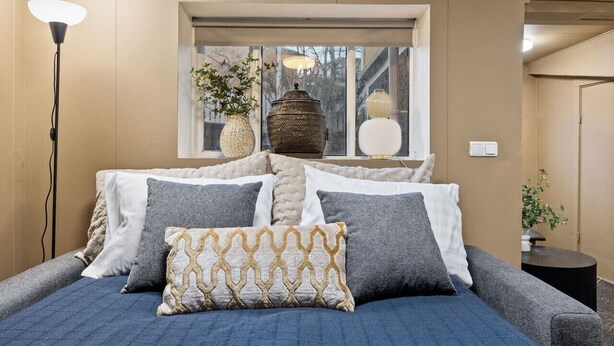 Ein Schlafzimmer mit Bett, Kissen, einer Stehlampe, einem Fenster mit Pflanzen und einer dekorativen Laterne.