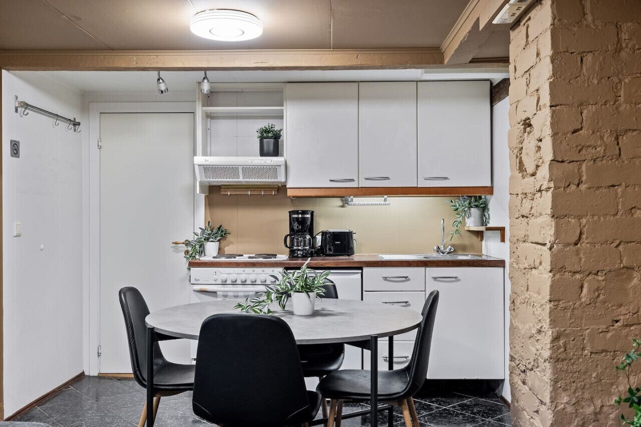 Cocina compacta con armarios blancos, estufa y cafetera. Hay una mesa redonda con sillas y plantas en macetas.