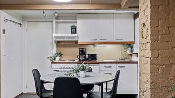 Cocina compacta con armarios blancos, estufa y cafetera. Hay una mesa redonda con sillas y plantas en macetas.