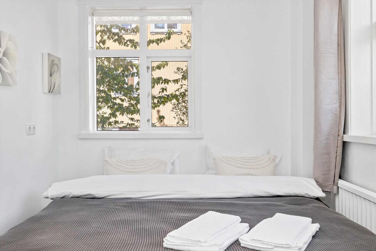 A bedroom with a bed, white curtains, and a window with a view of trees and buildings.