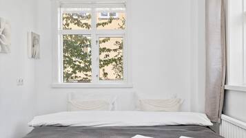 A bedroom with a bed, white curtains, and a window with a view of trees and buildings.