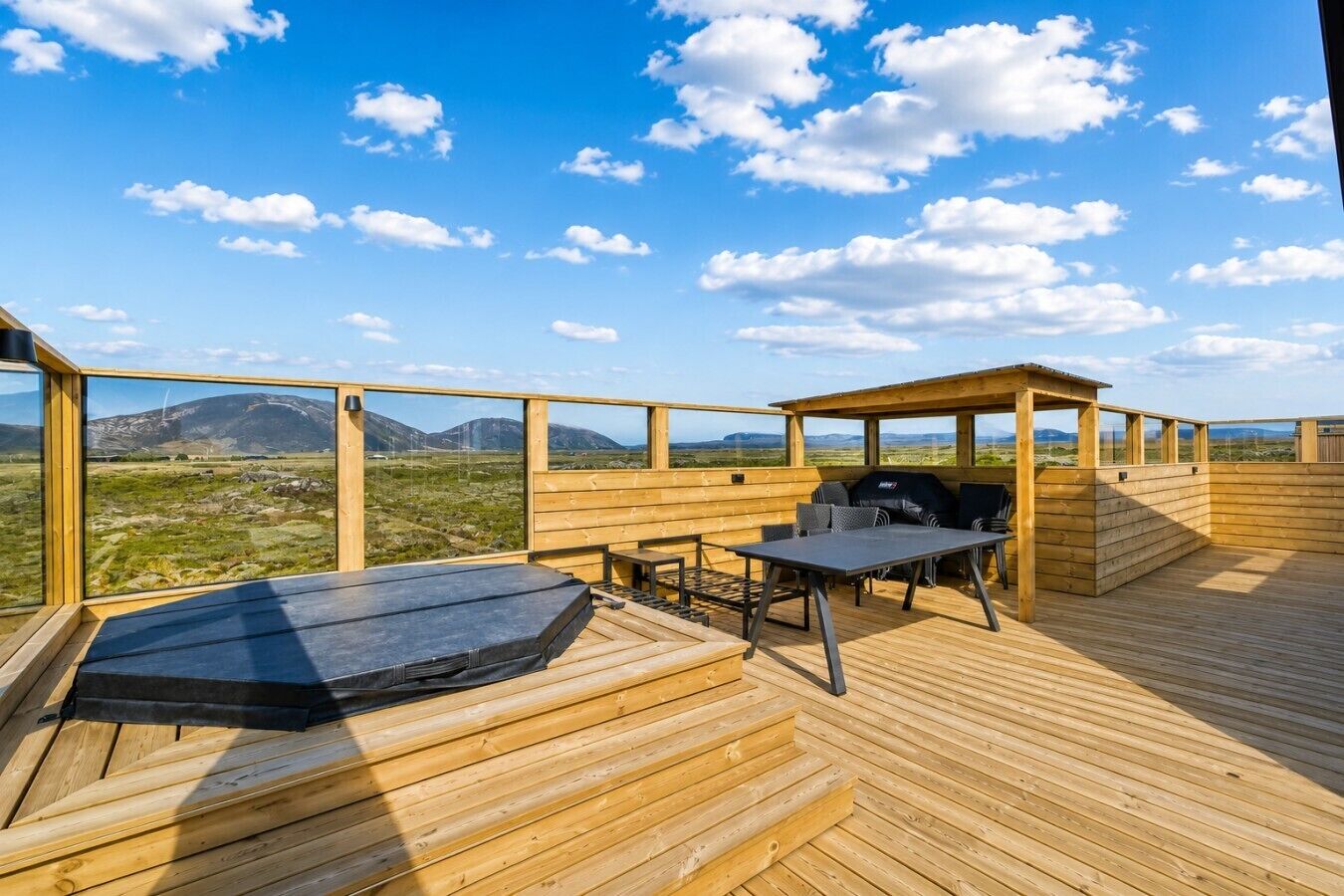 A wooden deck with a hot tub, grill, and table, overlooking a mountainous landscape.
