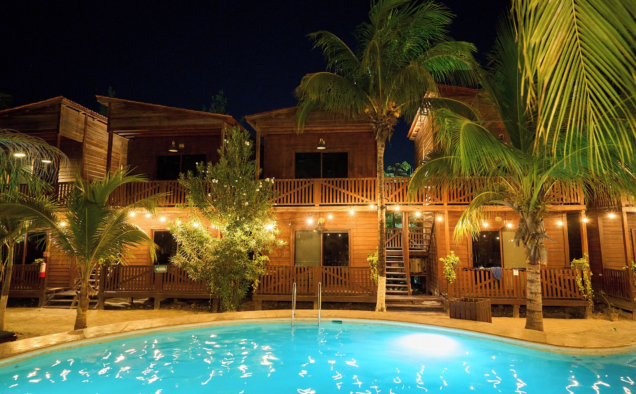 Un edificio de madera de dos pisos con piscina, rodeado de palmeras y luces colgantes.