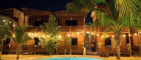 Un bâtiment en bois à deux étages, doté d’une piscine, entouré de palmiers et illuminé de guirlandes lumineuses.