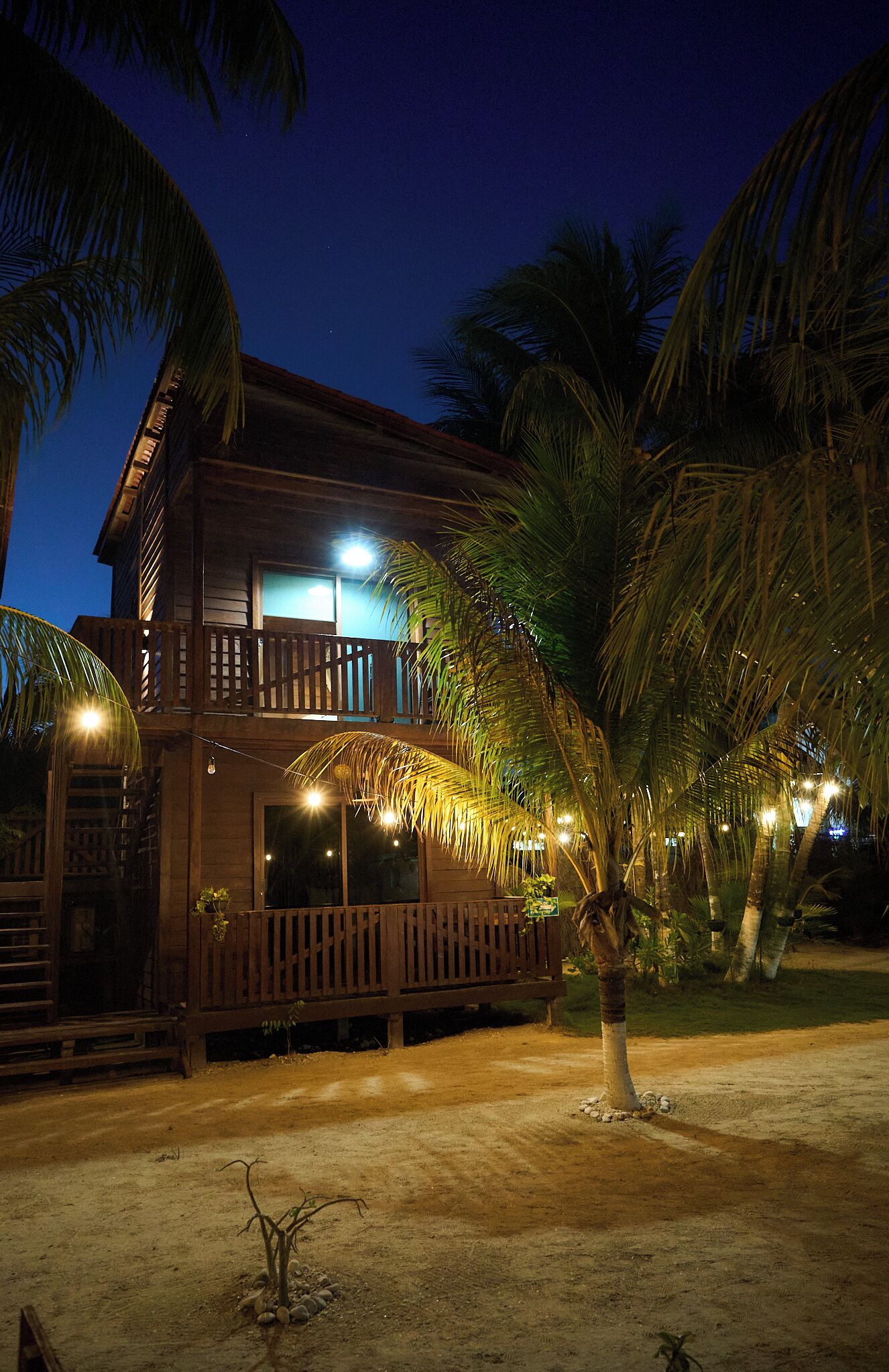 A two-story wooden building with a balcony, surrounded by palm trees and illuminated by string lights.