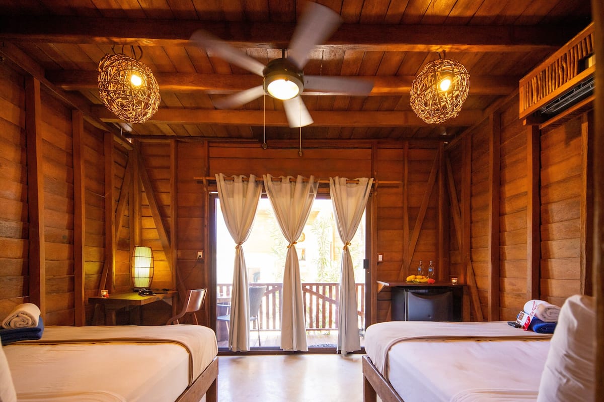 A wooden cabin room with two beds, a ceiling fan, and a balcony with curtains.