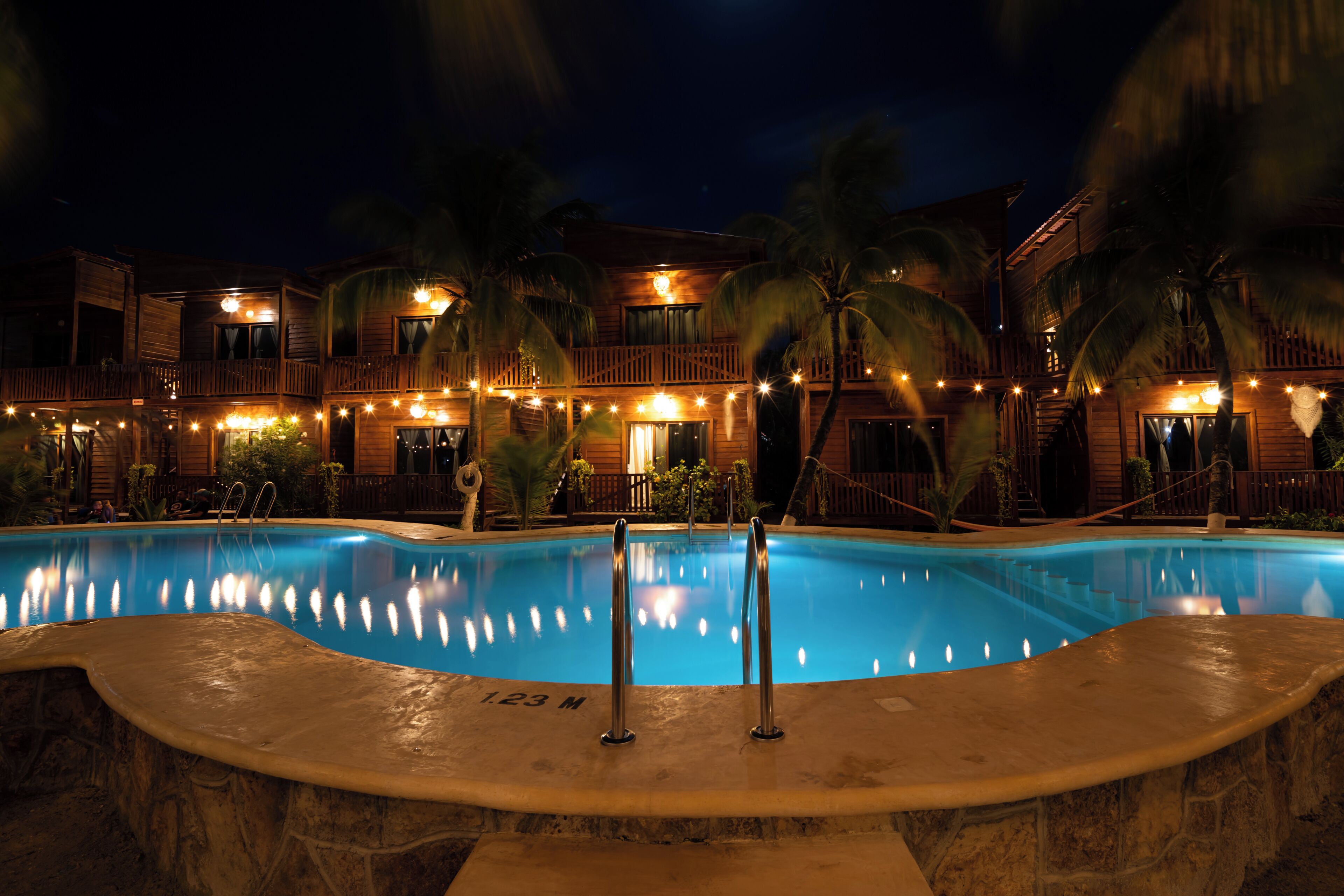 A swimming pool with a stone deck, surrounded by palm trees and a wooden building with balconies.
