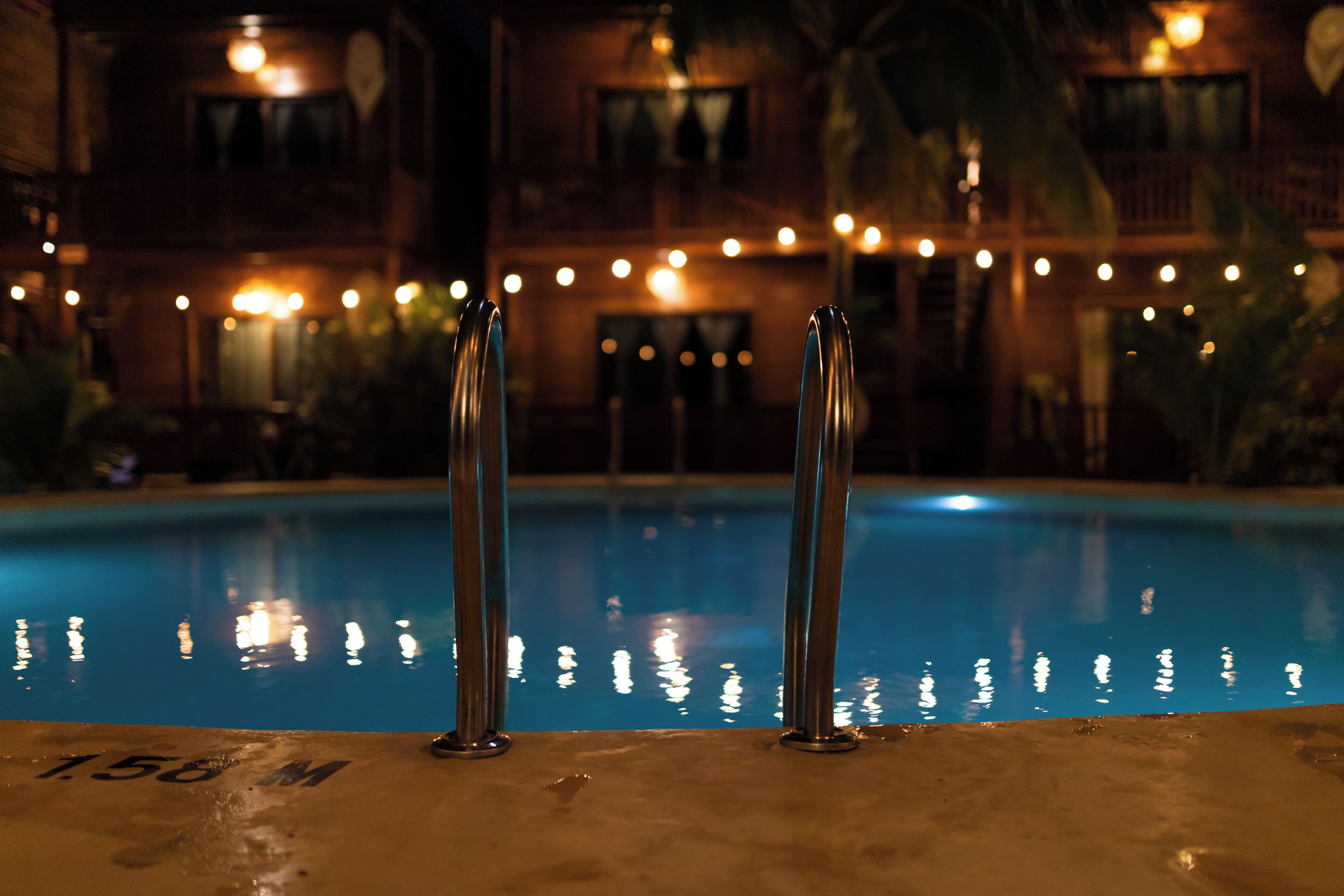 Une piscine avec des garde-corps en métal, le chiffre « 156 » inscrit sur le bord, et un bâtiment illuminé de guirlandes lumineuses en arrière-plan.