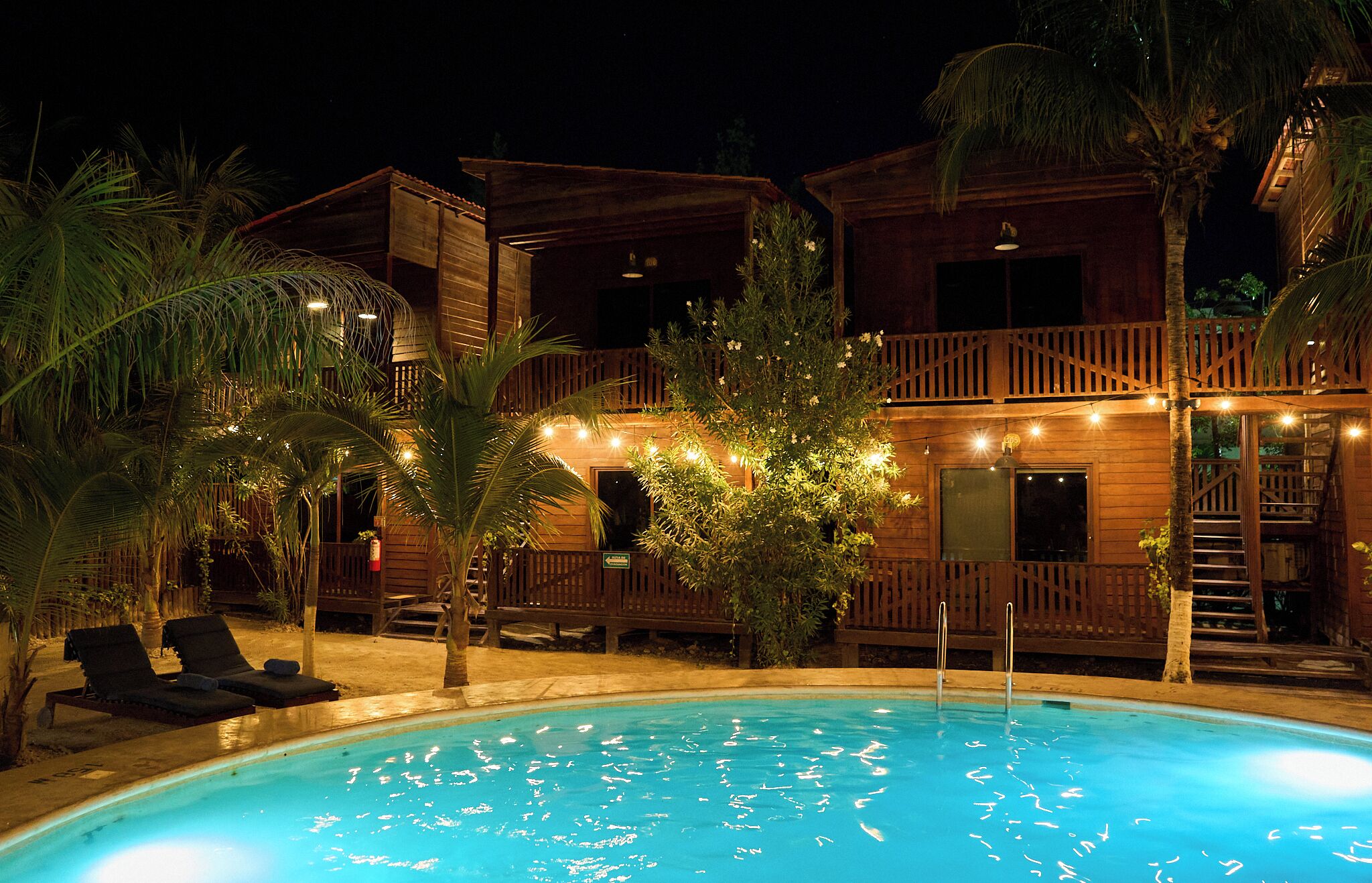 Un área junto a la piscina con un edificio de madera, sillas de descanso y una escalera que conduce a un nivel superior.