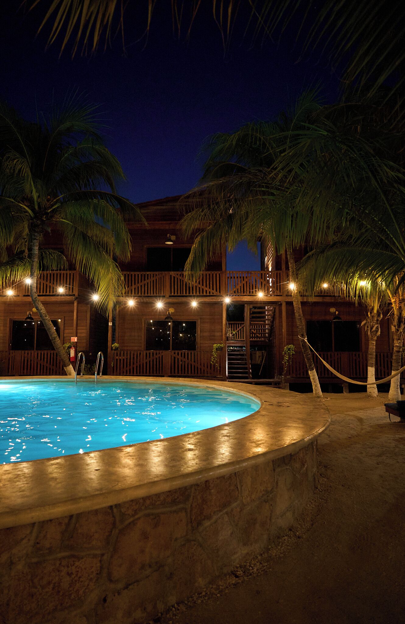 A pool area with a wooden deck, palm trees, and a building with balconies.