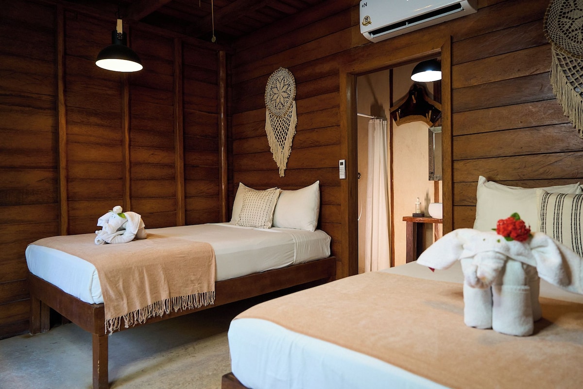 Habitación de madera con dos camas, aire acondicionado de pared y un adorno colgante de macramé.
