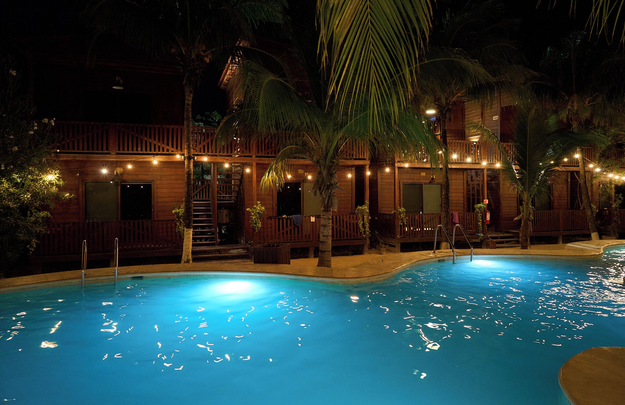 A nighttime pool area with wooden structures, palm trees, and string lights.