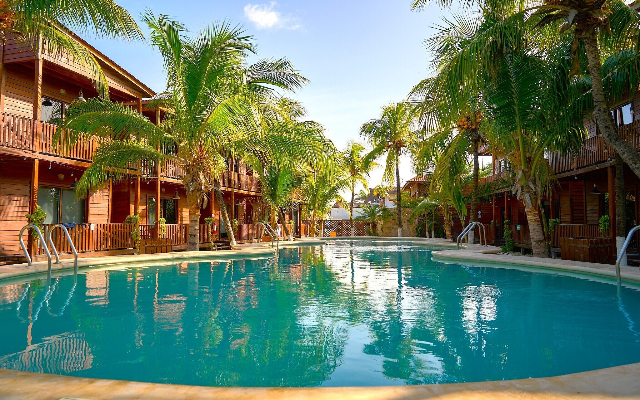 A swimming pool surrounded by wooden buildings with balconies, palm trees, and clear skies.