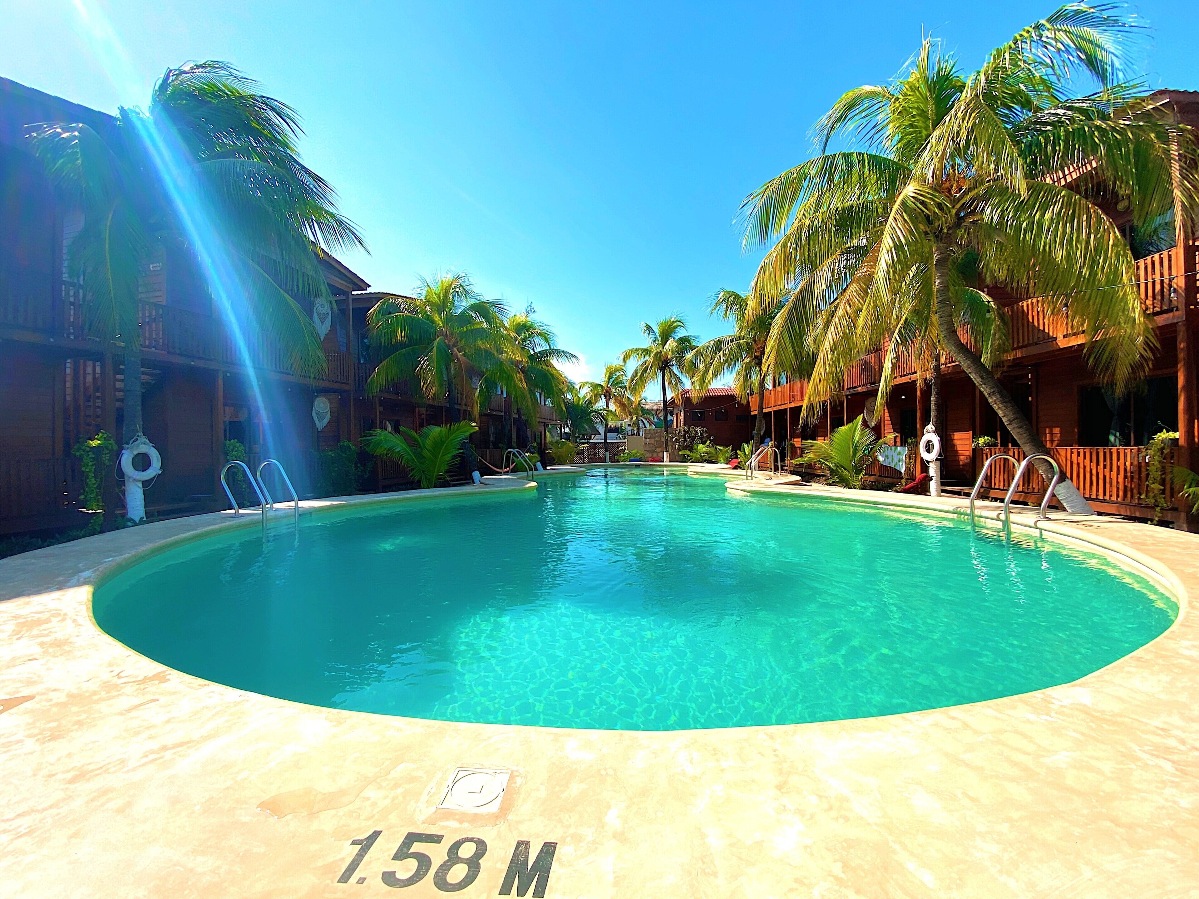 A large oval-shaped pool with clear blue water, surrounded by palm trees and wooden buildings.