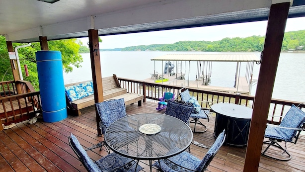 A wooden deck with a table and chairs, overlooking a lake with a dock and greenery in the background.