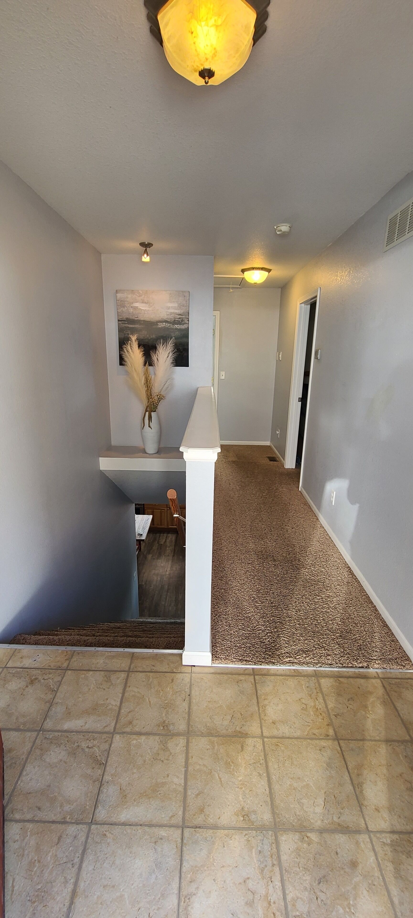 A hallway with a tiled floor, a white bench with a vase of pampas grass, and a painting on the wall.