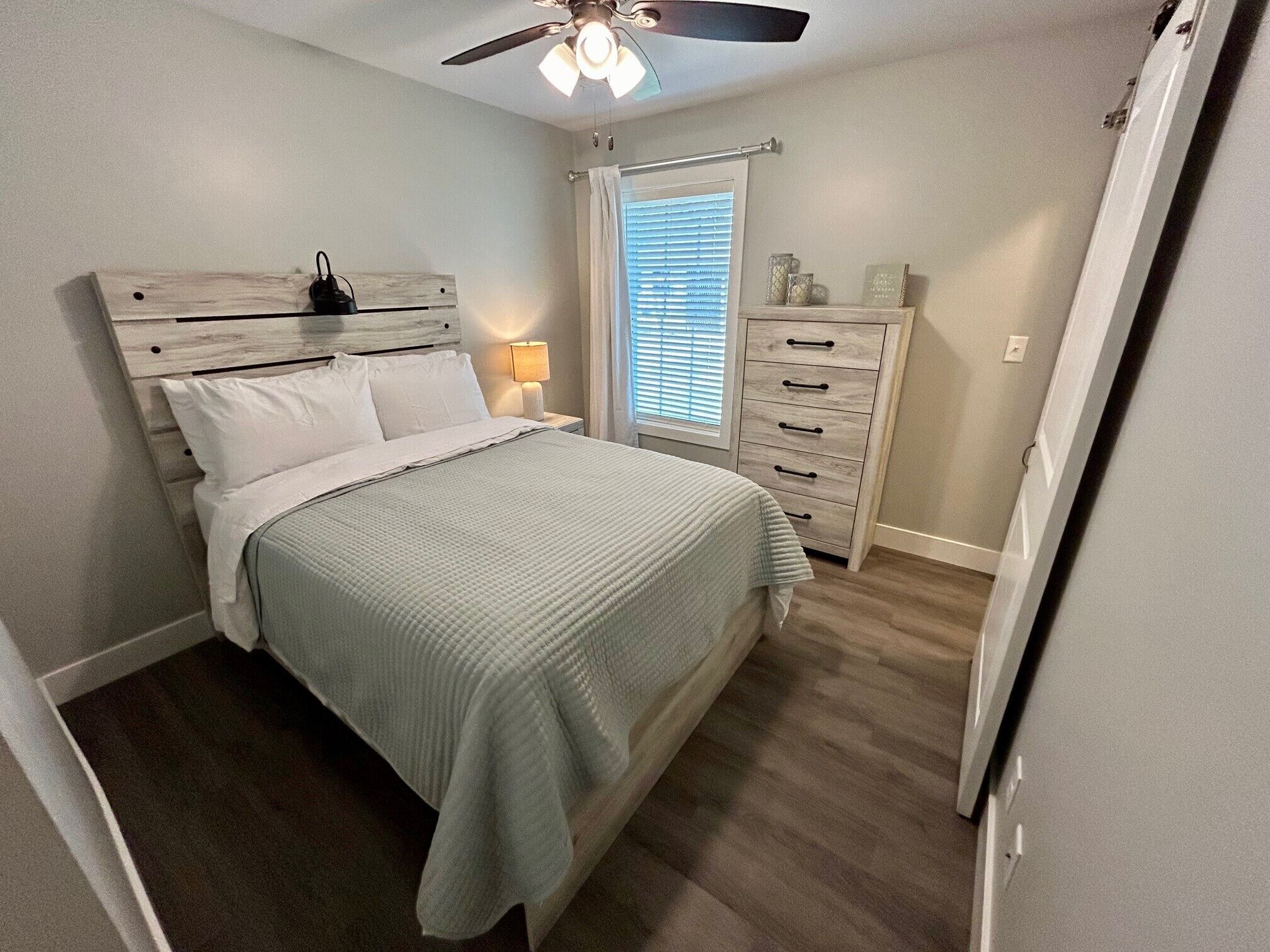 Une chambre à coucher comprenant un lit, une table de chevet, une commode, un ventilateur de plafond et une fenêtre avec des stores.