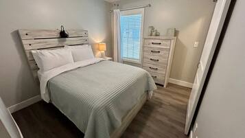 Une chambre à coucher comprenant un lit, une table de chevet, une commode, un ventilateur de plafond et une fenêtre avec des stores.